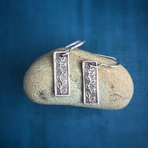 Silver Persist Earrings on a stone on a blue background. Earrings are small rectangles with vines. Empowering jewelry Vermont. 