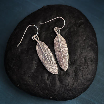 Two Silver Sage Leaf Earrings on a black stone sitting on a blue table.