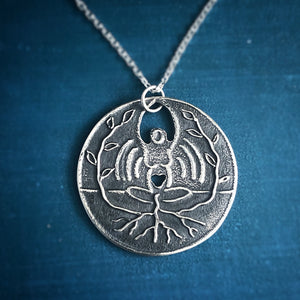 Silver Pregnancy Loss Miscarriage Necklace. Silver Necklace is woman sitting crosslegged with a heart cut out of her belly. Her arms are up. Roots comes down from the heart and into the ground, where new plants emerge and surround her. She is radiating a rainbow. 