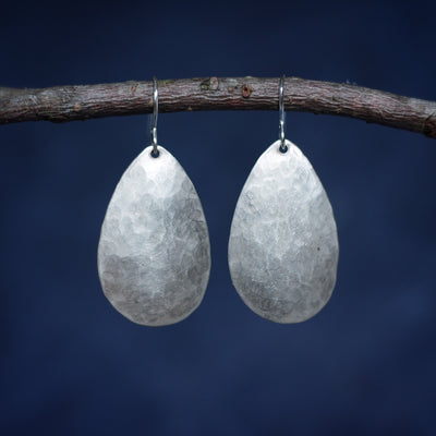 Grounded Earrings are egg shaped hammered silver earrings hanging on branch blue background empowering jewelry handmade in Vermont
