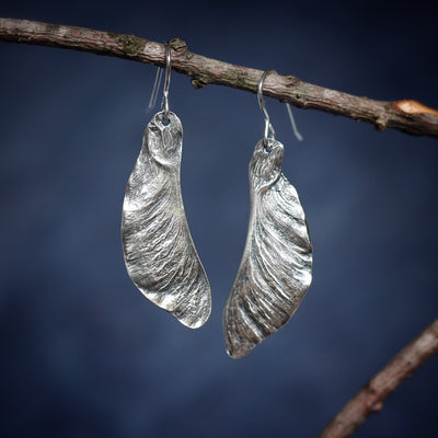 Silver Samara Maple Seed Earrings hanging on branch on blue background. Silver Maple Seed Jewelry handmade in Vermont