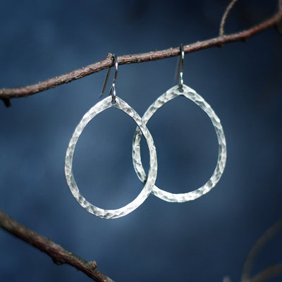 Embraced Earrings Silver hammered teardrop hoop earrings hanging on branch blue background Vermont empowering jewelry 