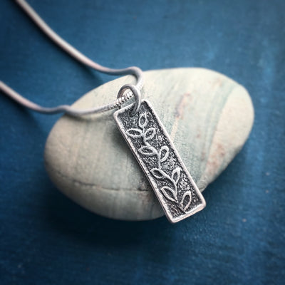 Silver Persist Necklace on a stone on a blue background. Necklace is a small rectangles with vines. Empowering jewelry handcrafted Vermont.
