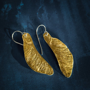 Gold Samara Maple Seed Earrings on a blue background. Seeds are handmade silver with 24 carat gold from Vermont maple seeds turned into earrings.