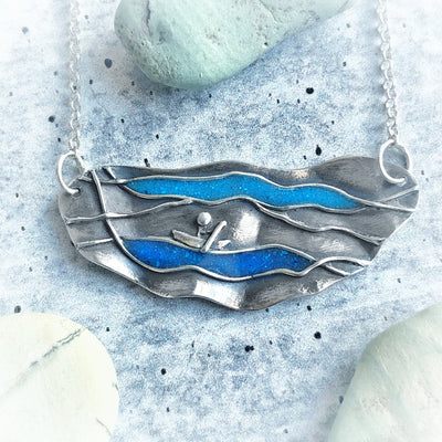 Silver Life's Currents Necklace on gray background. Silver necklaces has wavy edges with two blue turquoise currents and a small silver person on a canoe. Meaningful pendants, healing jewelry, empowering jewelry handmade in Vermont. 
