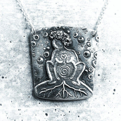  Silver Rooted Mama Necklace on cement background. Necklace has woman with curly hair squatting giving birth with a spiral in her belly and roots coming out of her. Birthing necklace and birthing jewelry gift for midwife postpartum.