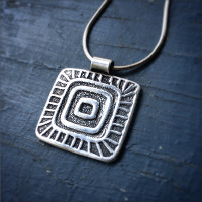 Silver Samay Pendant on Blue background. Square with concentric squares and lines surrounding it. Empowering jewelry Vermont